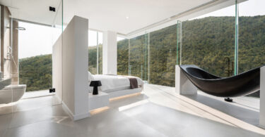bathroom behind bed, white bed, forest view bedroom, full height glass window, floor to floor glass window, black bathtub, counter basin,