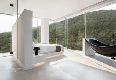 bathroom behind bed, white bed, forest view bedroom, full height glass window, floor to floor glass window, black bathtub, counter basin,