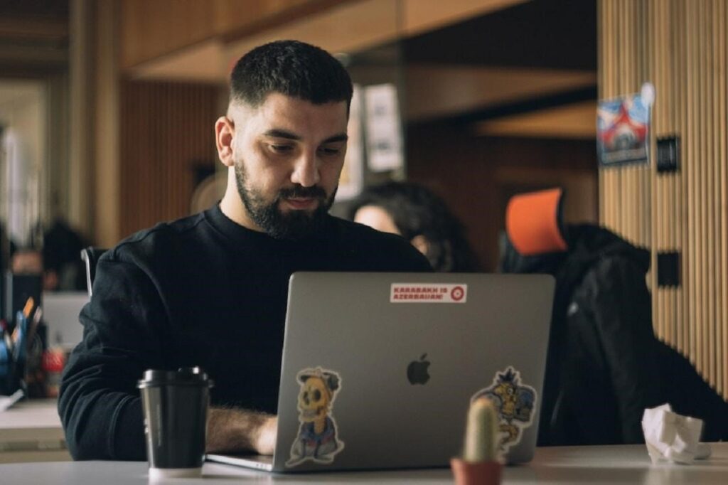 A man sitting at a work desk using his laptop with his coffee cup placed next to it.